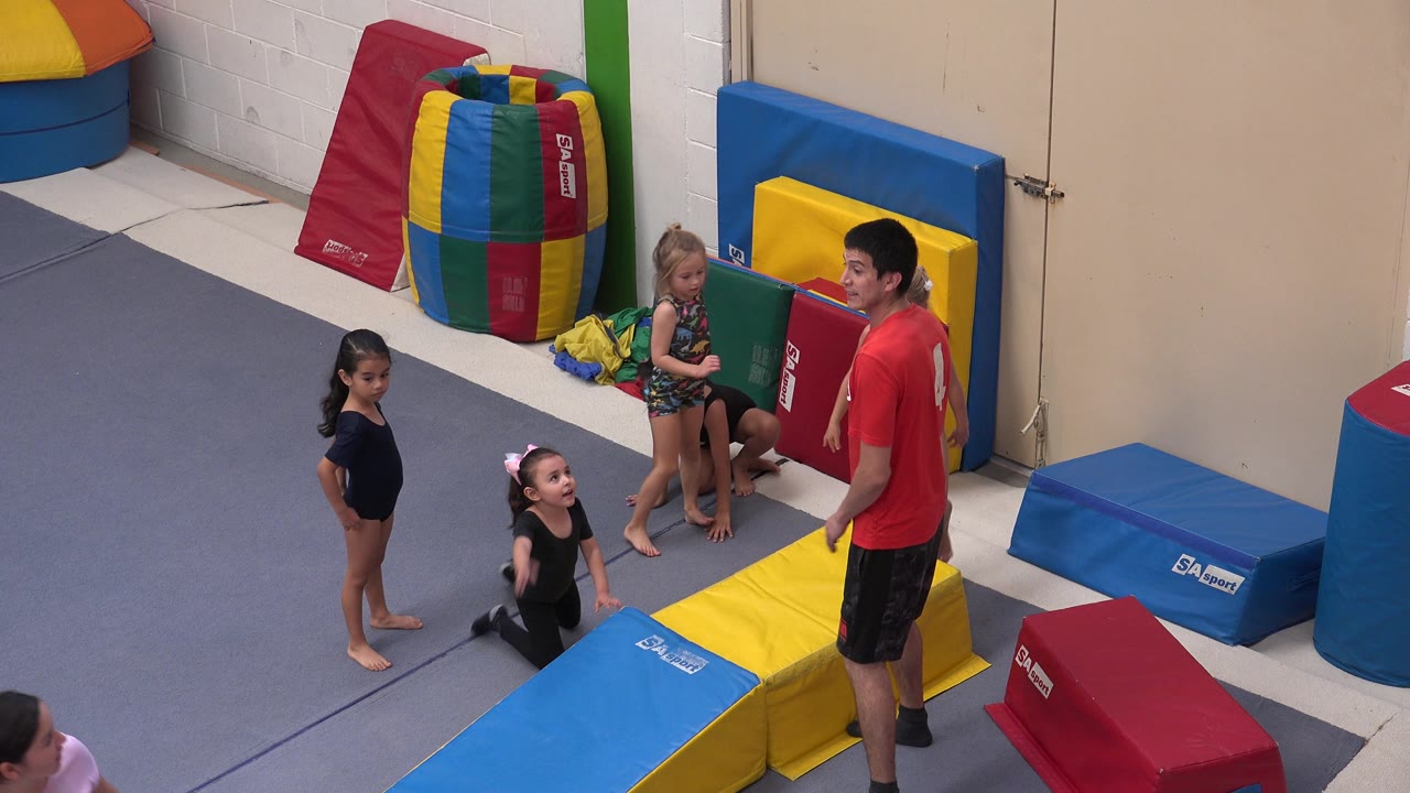 In Cabo San Lucas, Mexico, two children stand, one child kneels, and another child sits near the wall as the instructor faces the group beside padded gym equipment.