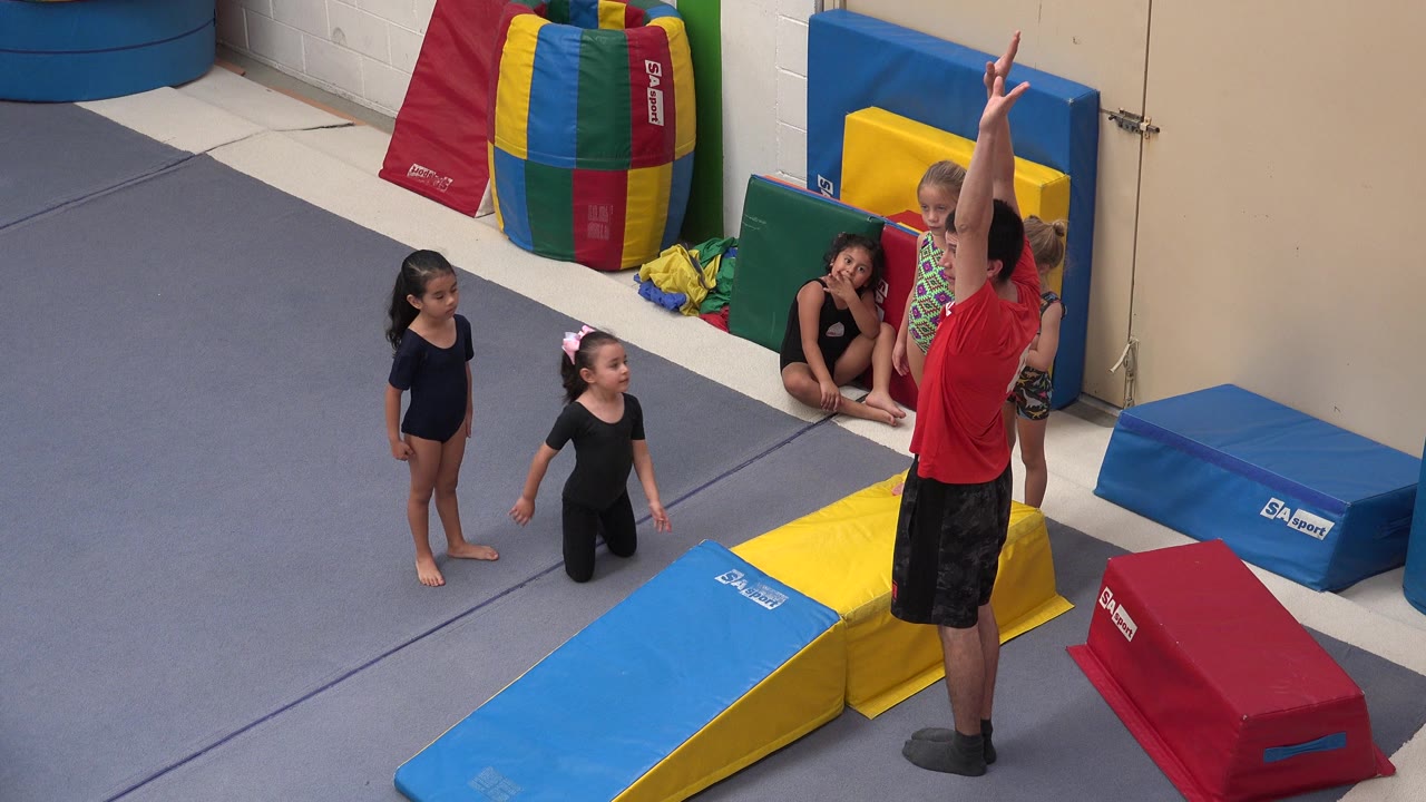 Girls watch a gymnastics instructor beside foam mats.