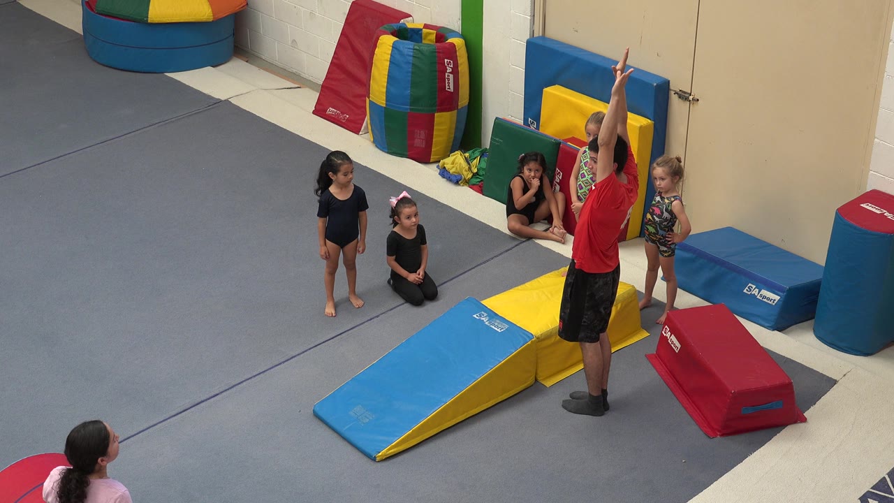 Instructor in red shirt raises both arms near children and foam gym mats.