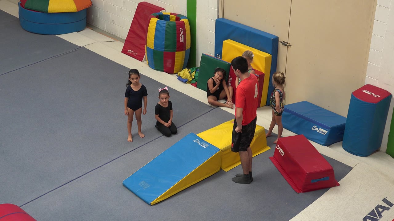 Instructor in red shirt stands near children and padded gym equipment.
