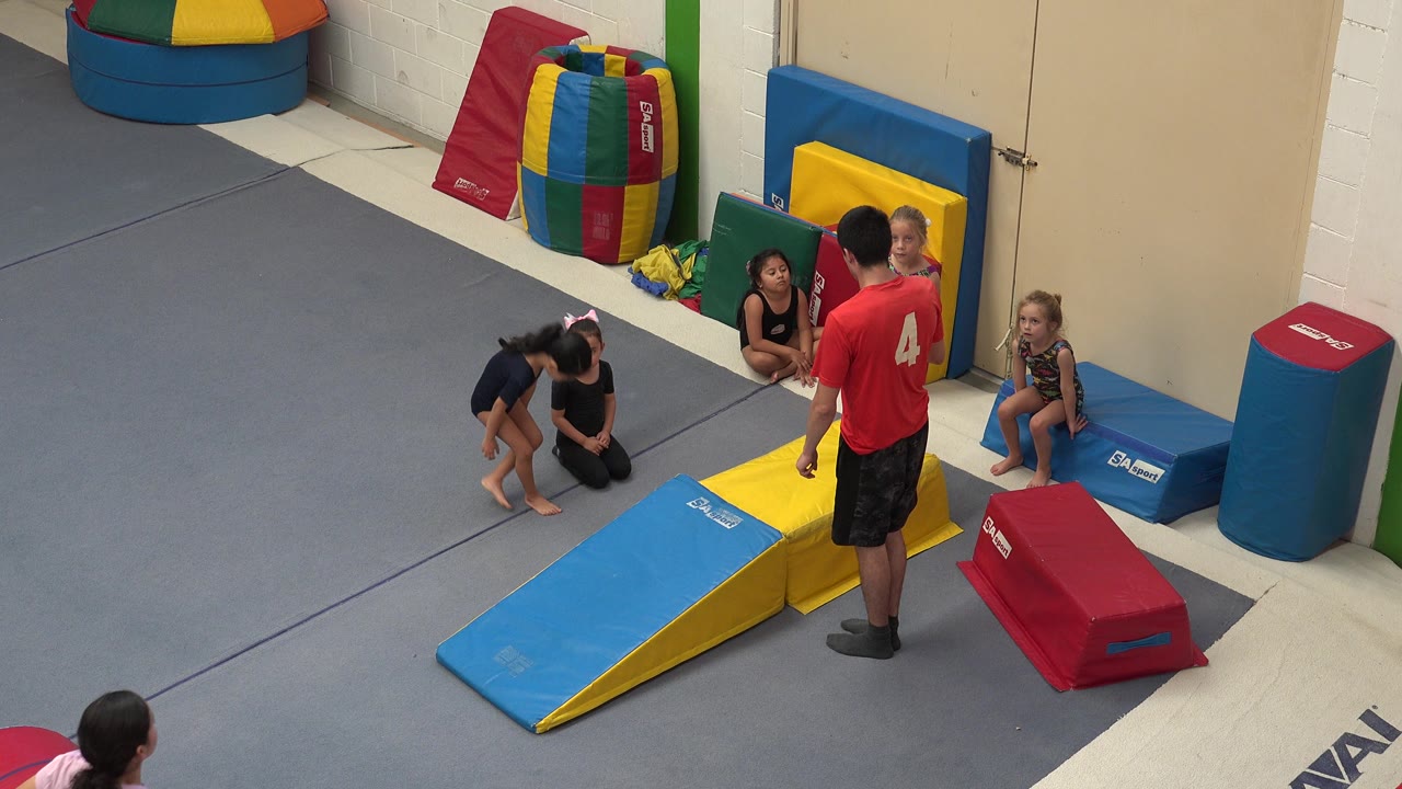 In Cabo San Lucas, Mexico, children sit and crouch on the padded floor as the coach stands near the yellow block and blue ramp in the play area.