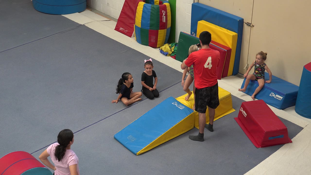 Instructor stands beside children and padded gymnastics mats in an indoor gym.