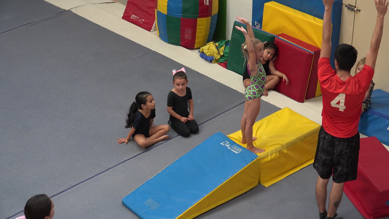 Coach in red raises his arms as a young gymnast stands on foam mats.