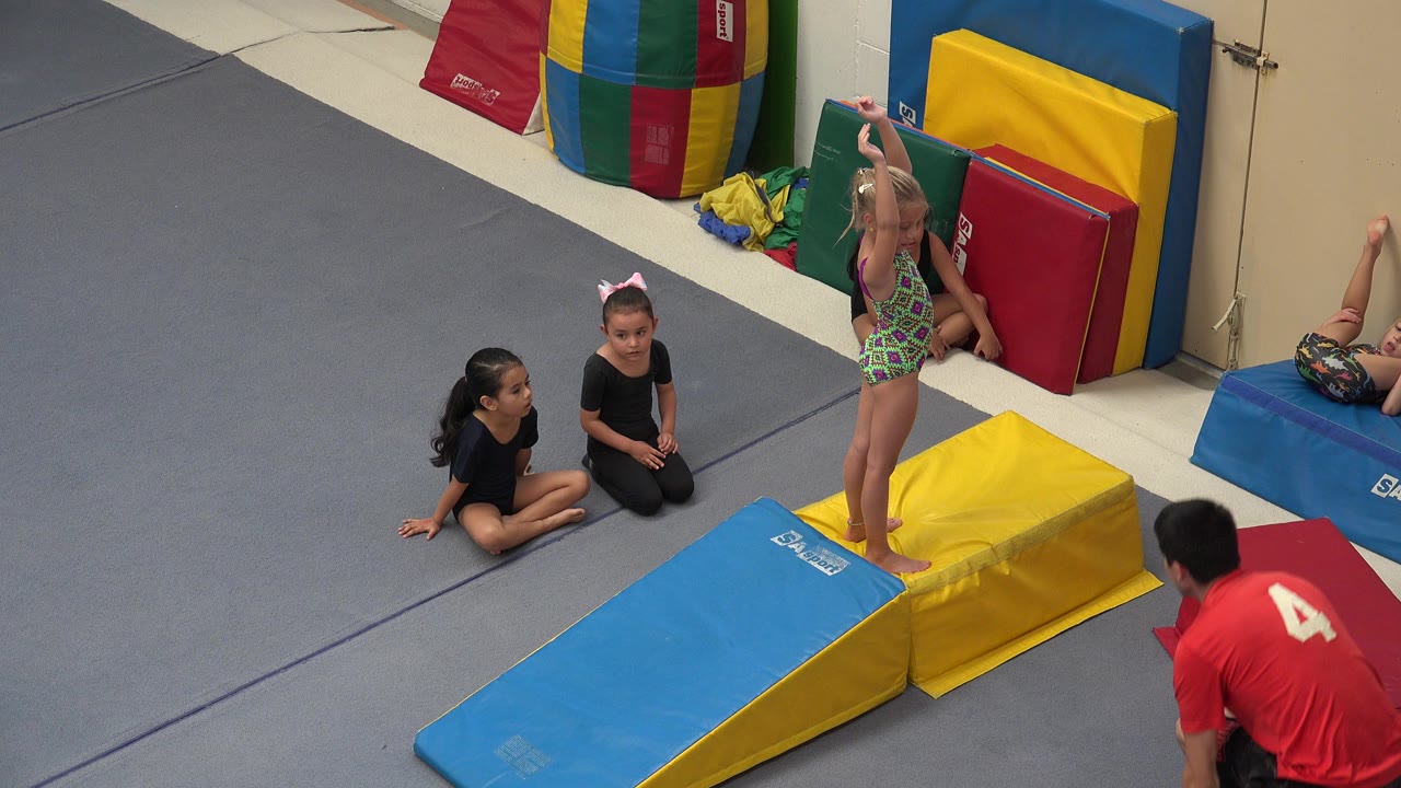 Young girl standing on a blue and yellow gym mat with seated girls nearby.