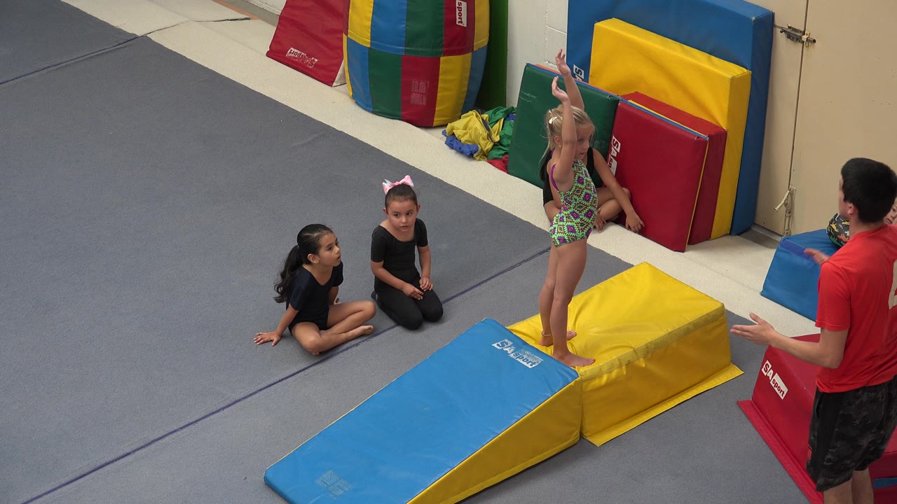 Instructor stands near a young girl on a padded gym ramp.