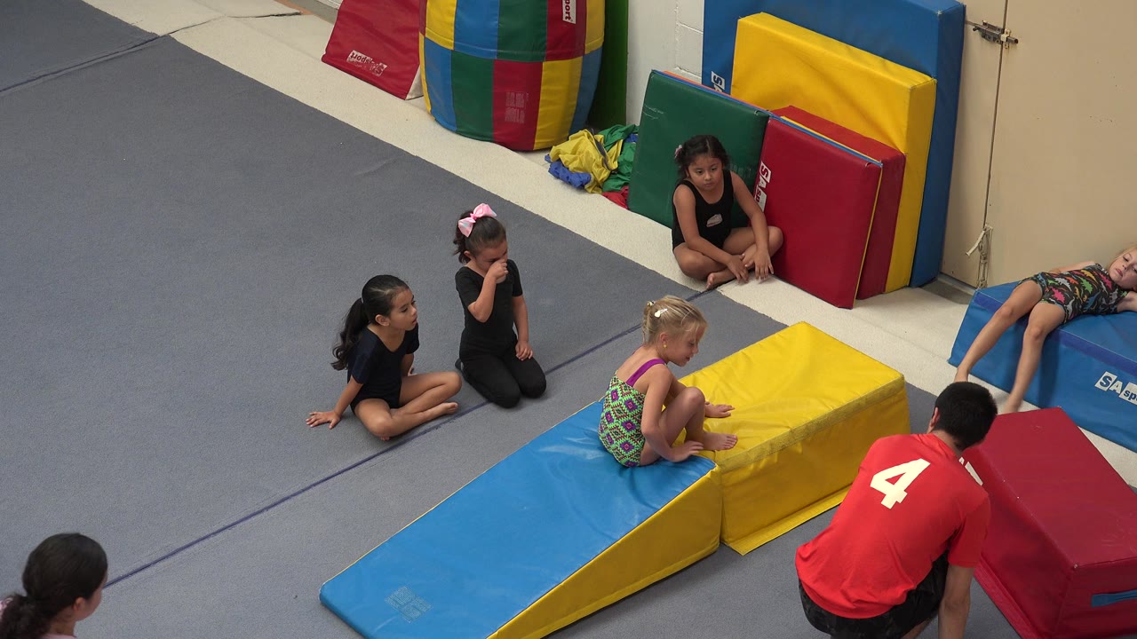In Cabo San Lucas, Mexico, several children sit near blue, yellow, red, and green foam tumbling mats while a girl crouches on a padded wedge and a person in a red shirt kneels nearby.