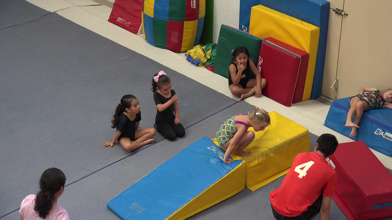 Children watch a tumbling station with foam mats and stacked pads.