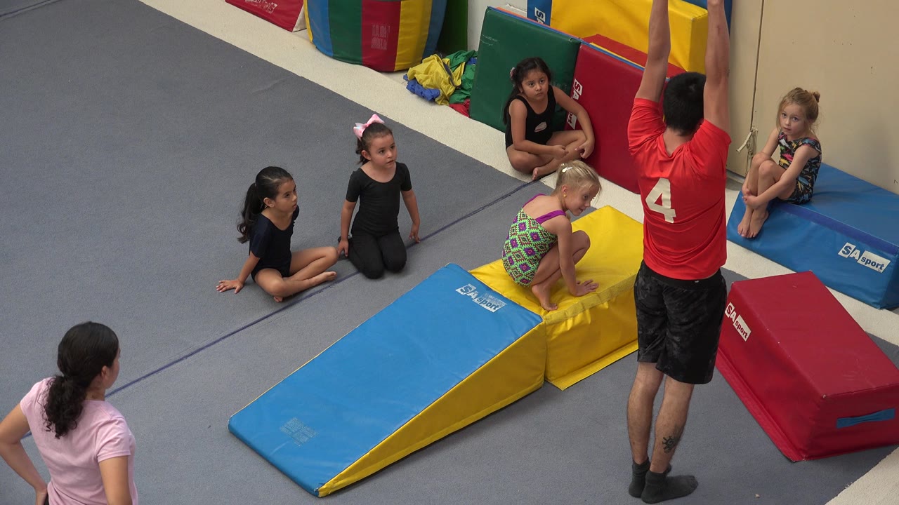In Cabo San Lucas, Mexico, a coach in a red shirt raises his arms beside children seated around yellow, blue, and red gymnastics pads at the start of the clip.