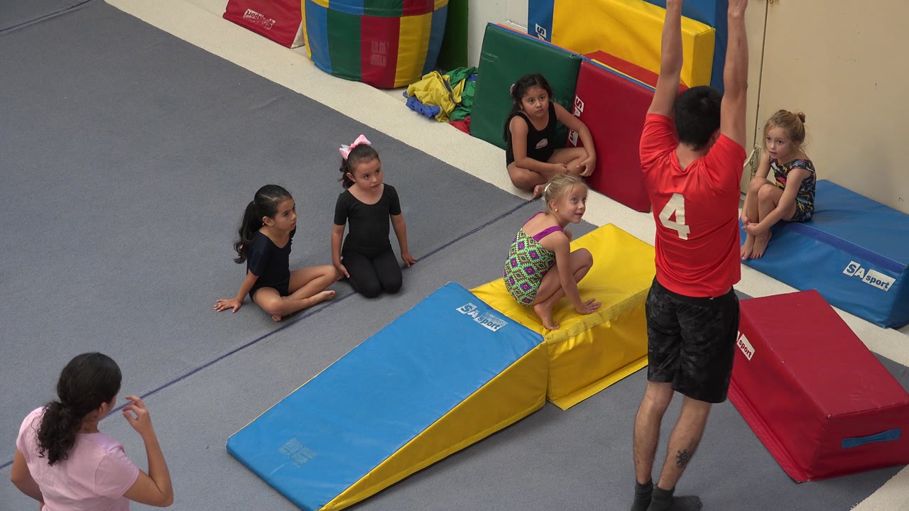 Coach stands beside padded equipment as children wait on the gym floor.