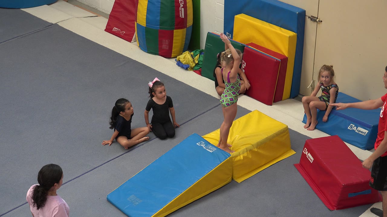Young girl in a green leotard stands on a yellow padded block in a gym.