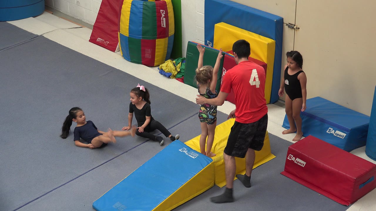 In Cabo San Lucas, Mexico, children sit and stand on gray gym flooring while an adult instructor in a red shirt stands near blue and yellow padded training blocks.