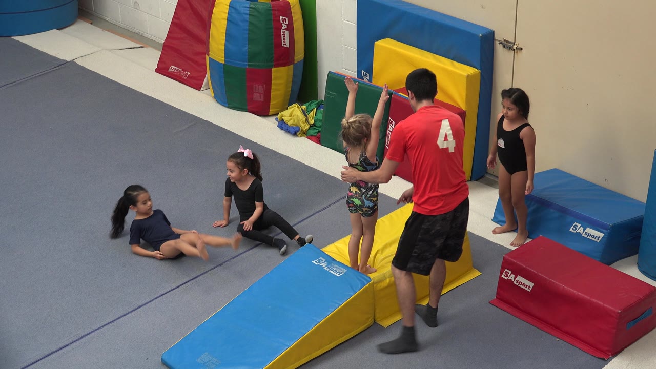 In Cabo San Lucas, Mexico, an instructor faces a child holding her arms up beside yellow and blue mats while other children sit or stand nearby in the gym.