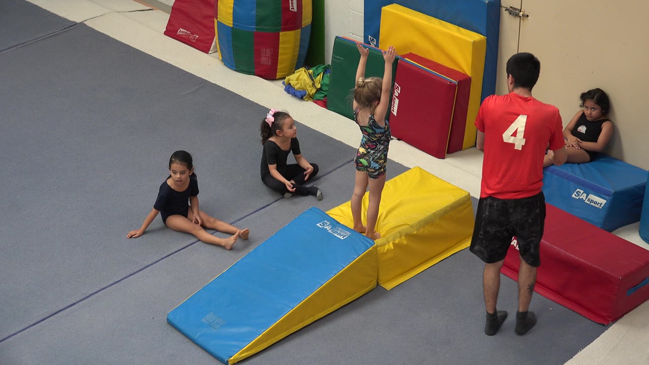 In Cabo San Lucas, Mexico, young girls gather around blue and yellow foam gymnastics mats while a coach in a red shirt stands nearby.