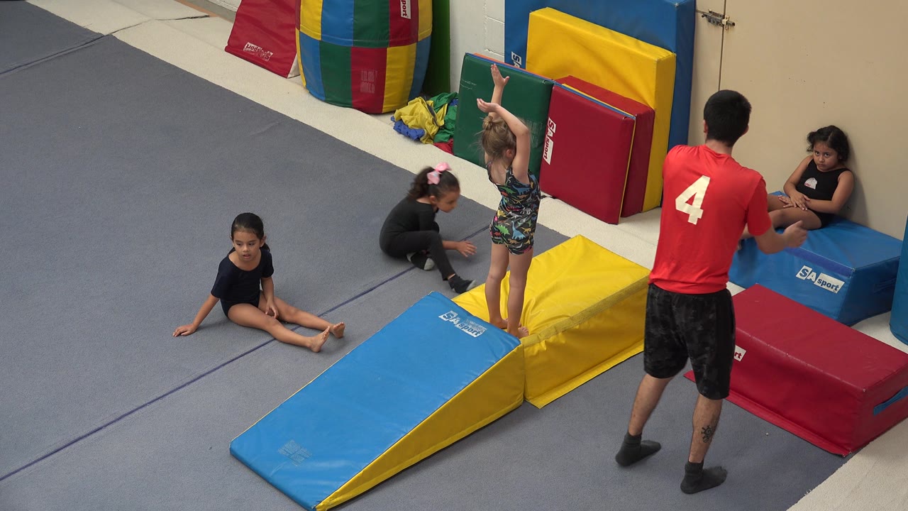 Coach gestures near seated girls and colorful gym pads.