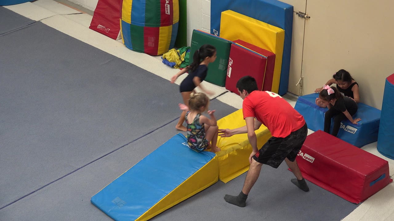 At 0:00 in Cabo San Lucas, Mexico, a male instructor in a red shirt leans toward a young child seated on a yellow foam wedge while other children wait near padded blocks.
