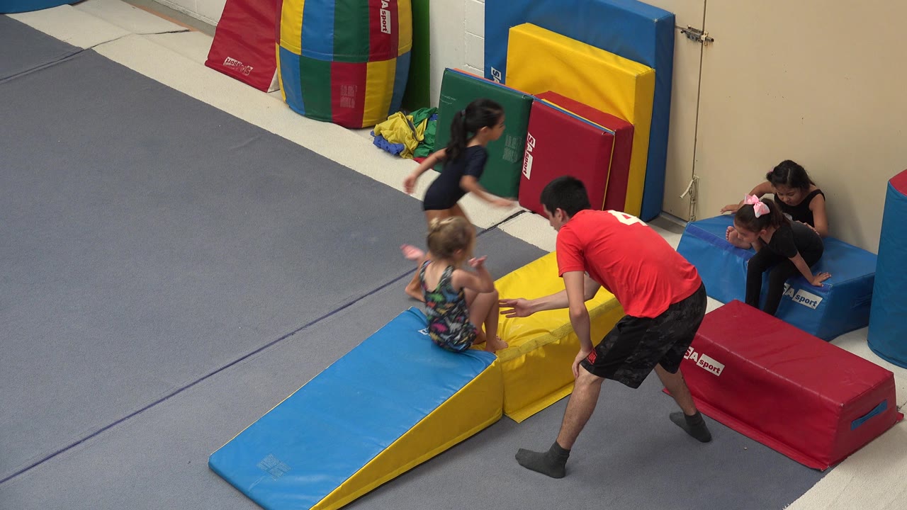 At 0:01 in Cabo San Lucas, Mexico, the child leans forward on the yellow wedge as the instructor stands close and other children remain along the padded wall area.
