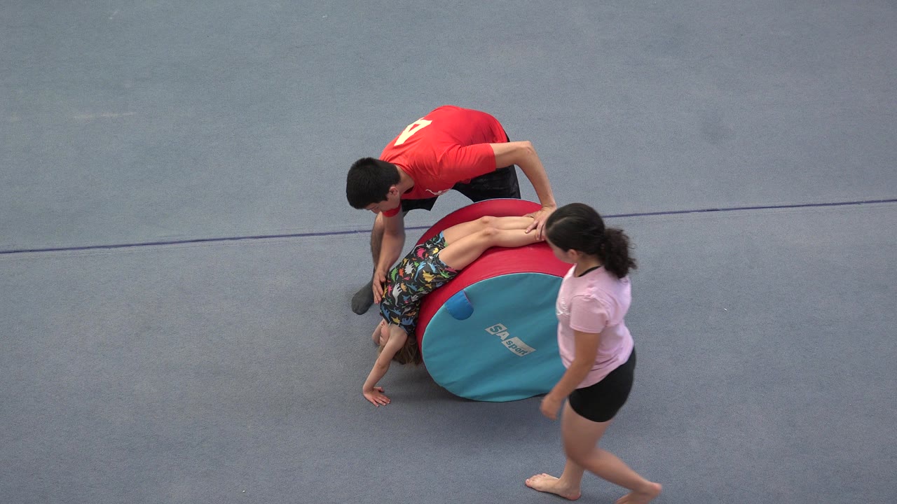 Adults assist a child beside a red and teal padded gym cylinder.