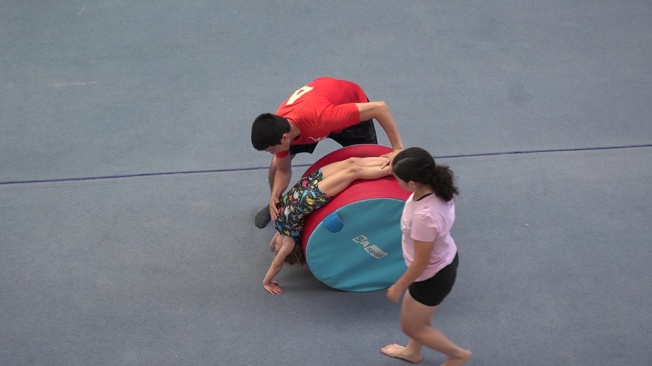 In Cabo San Lucas, Mexico, a child lies or leans across a red and teal padded cylinder while two adults stand close on the gray gym floor.