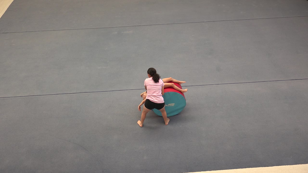Adult woman assists a girl on a foam cylinder in a gym.
