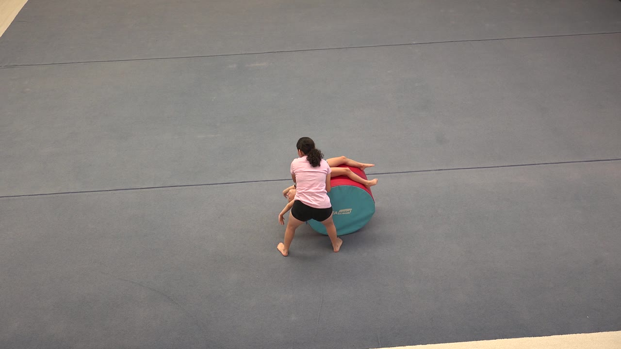 In Cabo San Lucas, the adult helper supports the young girl as her legs extend across the top of the red and teal cylinder on the gray mat.