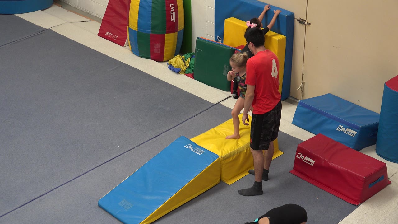 Trainer stands beside a child on yellow gym mats near a blue foam ramp.