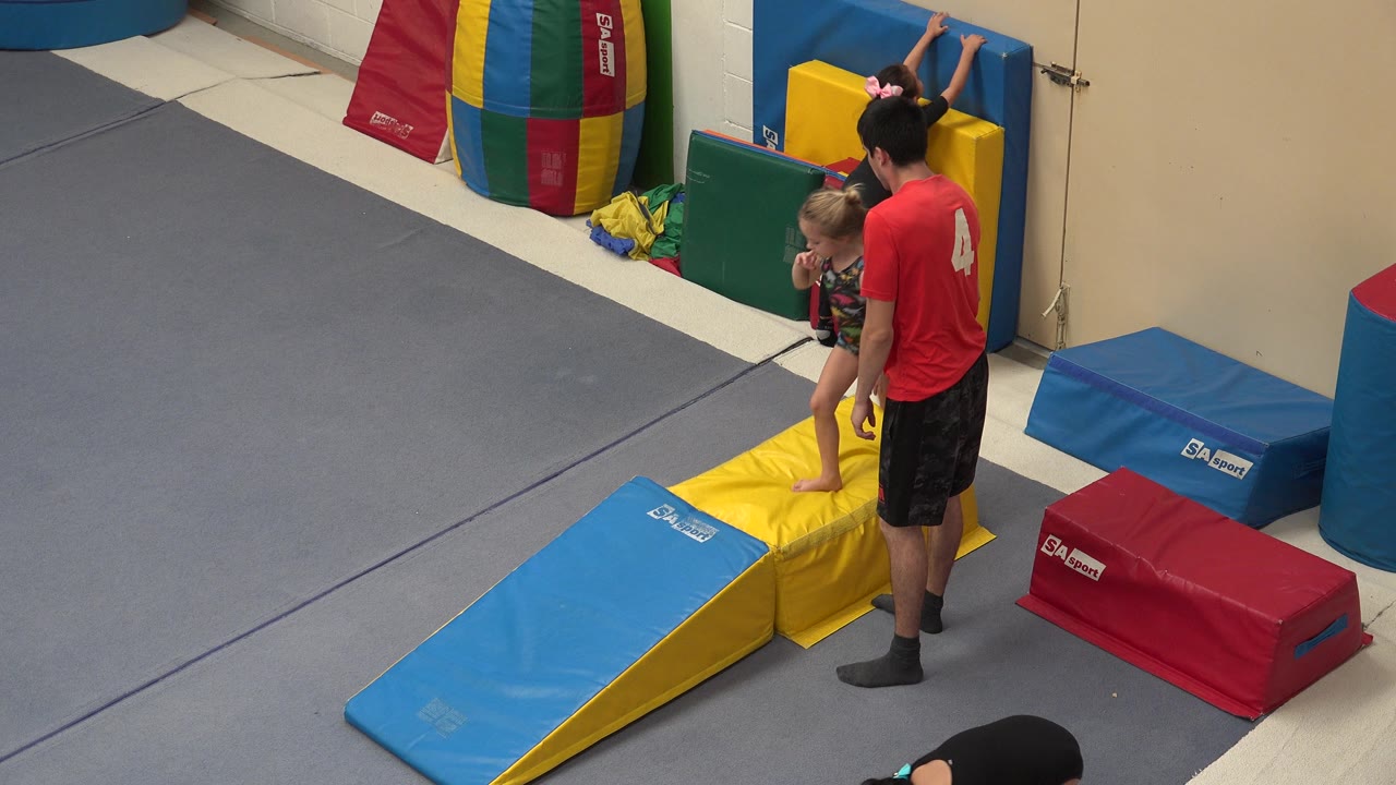 In Cabo San Lucas, Mexico, the trainer remains beside the child at the yellow mat station, with the blue ramp in the foreground and multicolor pads stacked against the wall.