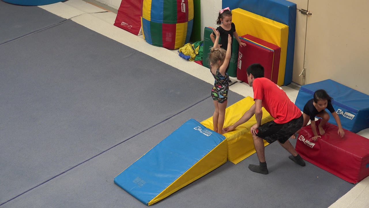 Instructor helps a child on padded tumbling mats while other children wait nearby.