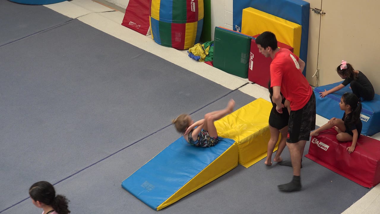 The girl is mid-somersault on the padded incline as the instructor watches from the side and two other girls sit or kneel near the red mat.
