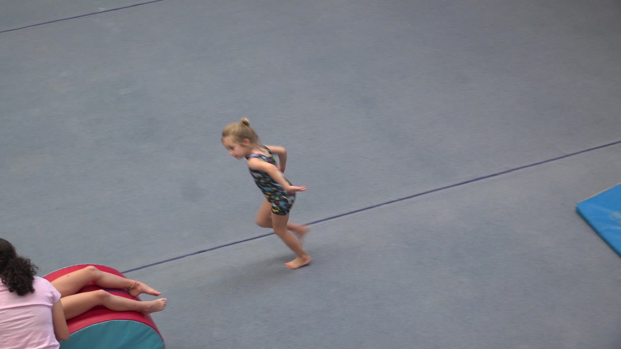 Young gymnast running on a blue gym floor near a coach and padded cylinder.