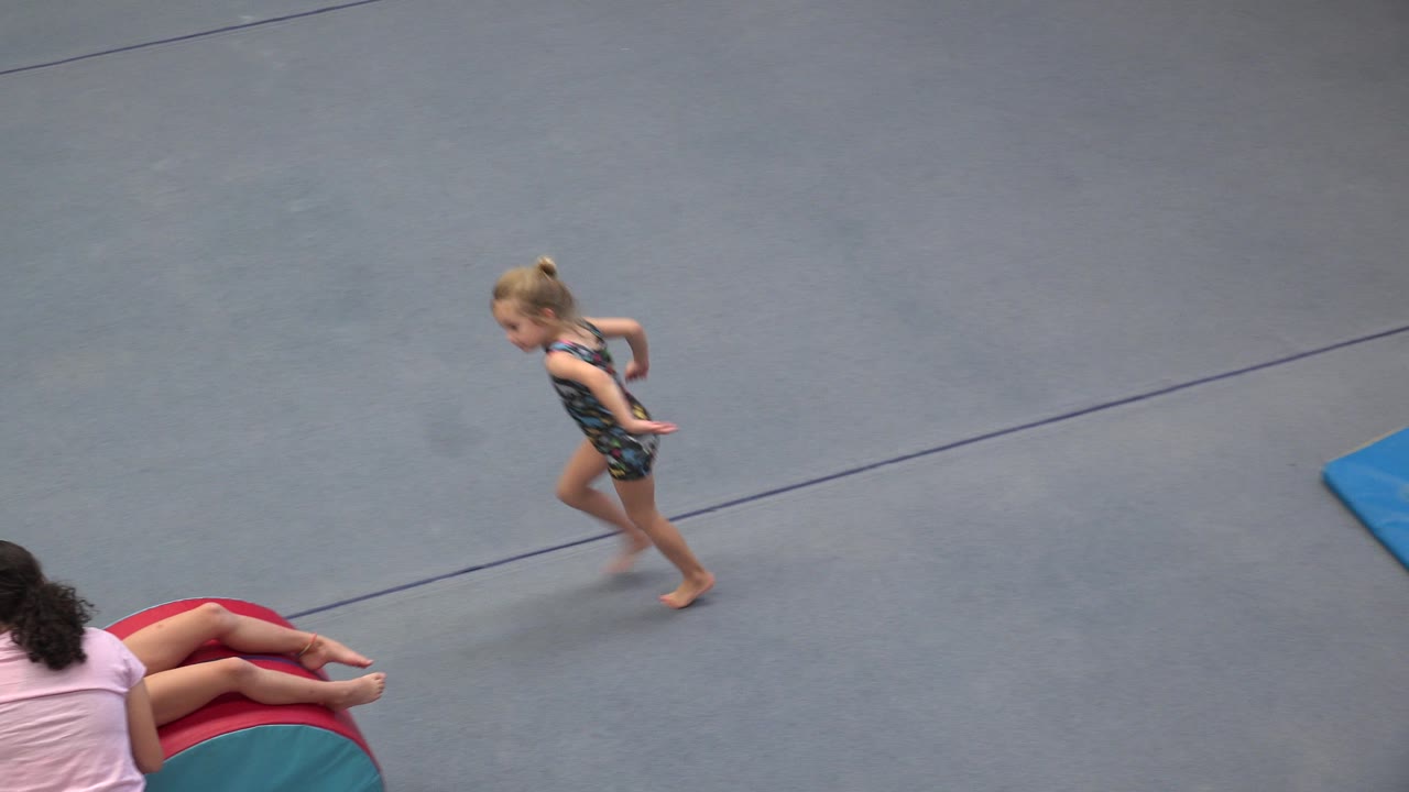 In Cabo San Lucas, Mexico, the young gymnast moves farther across the blue floor as the coach remains low beside the red and teal training cylinder.