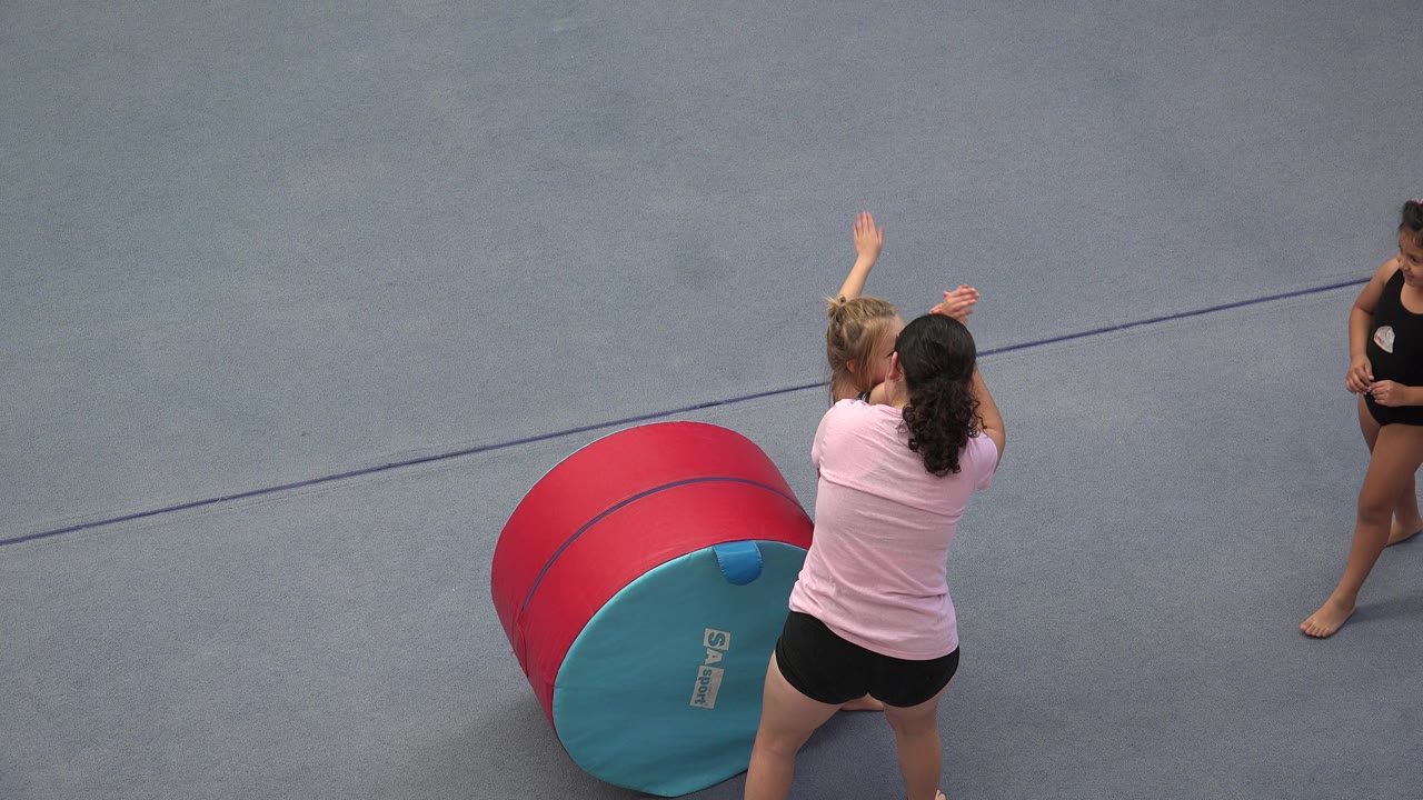 In Cabo San Lucas, Mexico, the instructor stands close to the girl as the child keeps both arms up next to the red and teal tumbling barrel.
