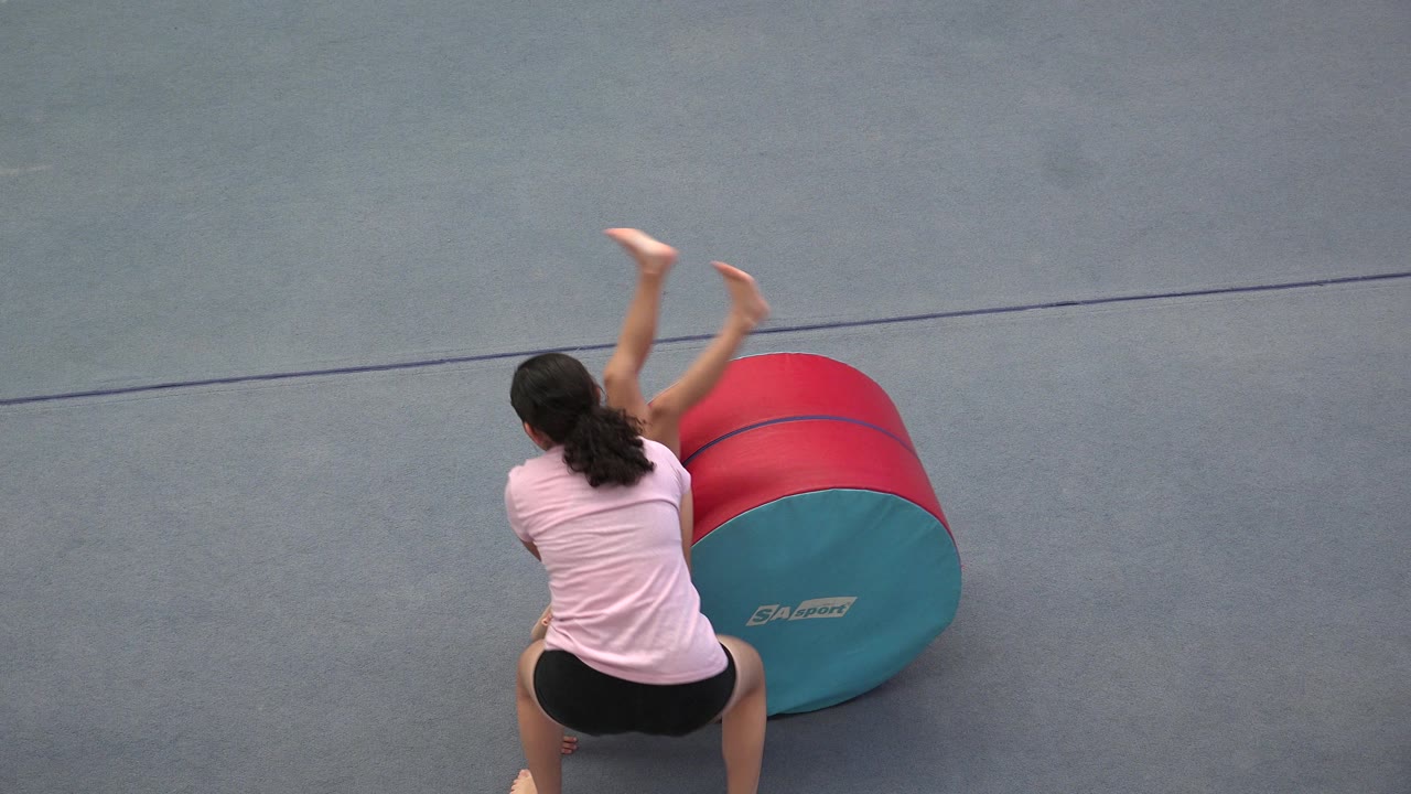 In Cabo San Lucas, Mexico, an adult instructor in a pink shirt stands near a red and teal training cylinder while a child finishes a tumble on a gray gym mat.