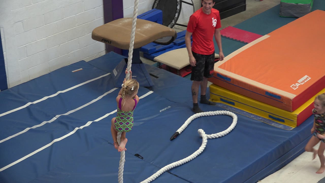 Toddler holding a hanging rope over blue gym mats near an instructor.