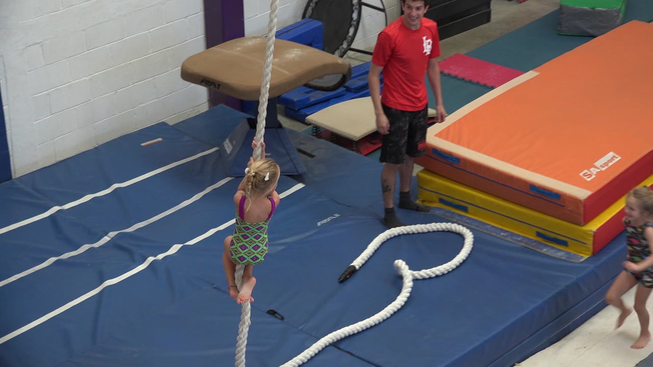 The toddler remains suspended on the rope in the Cabo San Lucas gym while the instructor watches and another child moves into the right side of the padded exercise area.