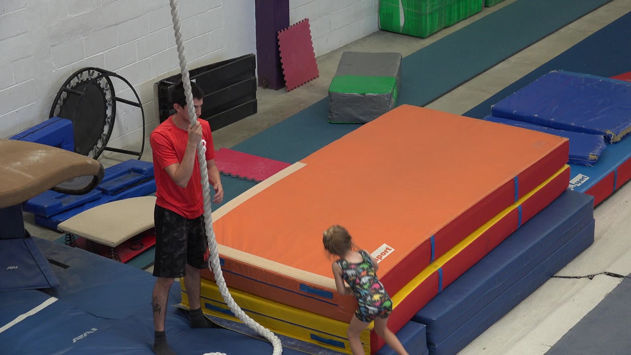 In Cabo San Lucas, a young girl in a patterned leotard approaches a thick white rope while an adult man in a red shirt stands beside orange and blue gym mats.