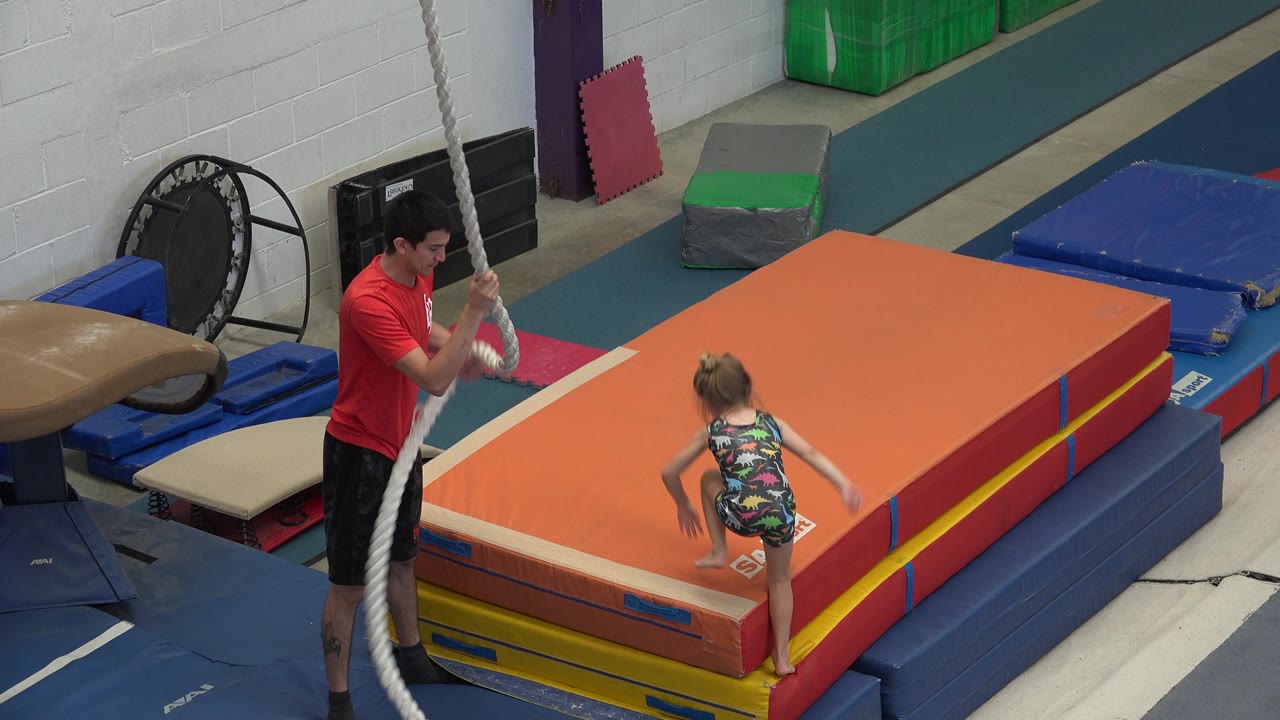 Girl climbs onto the orange mat near the rope and coach.