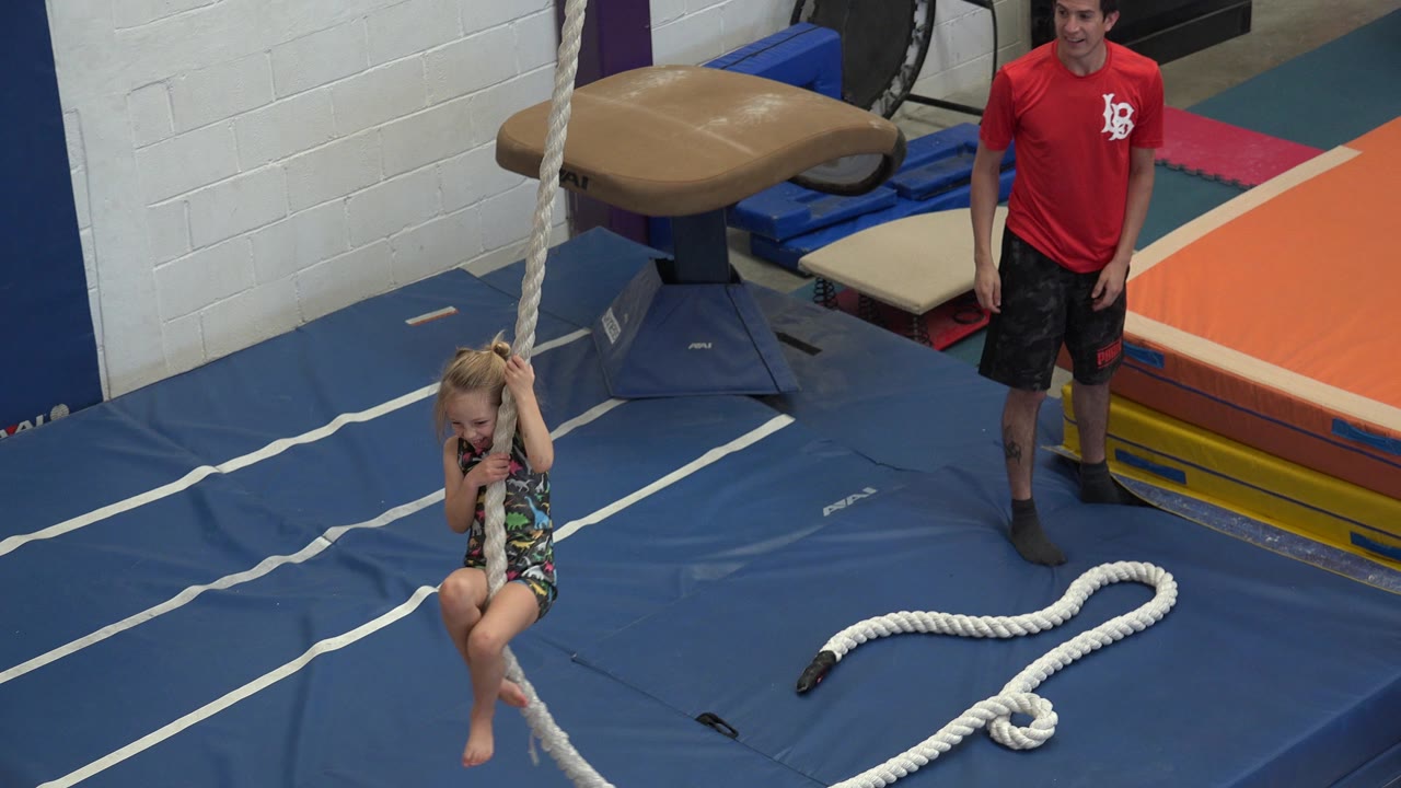 Young girl holds a hanging rope on blue gym mats as a trainer stands nearby.