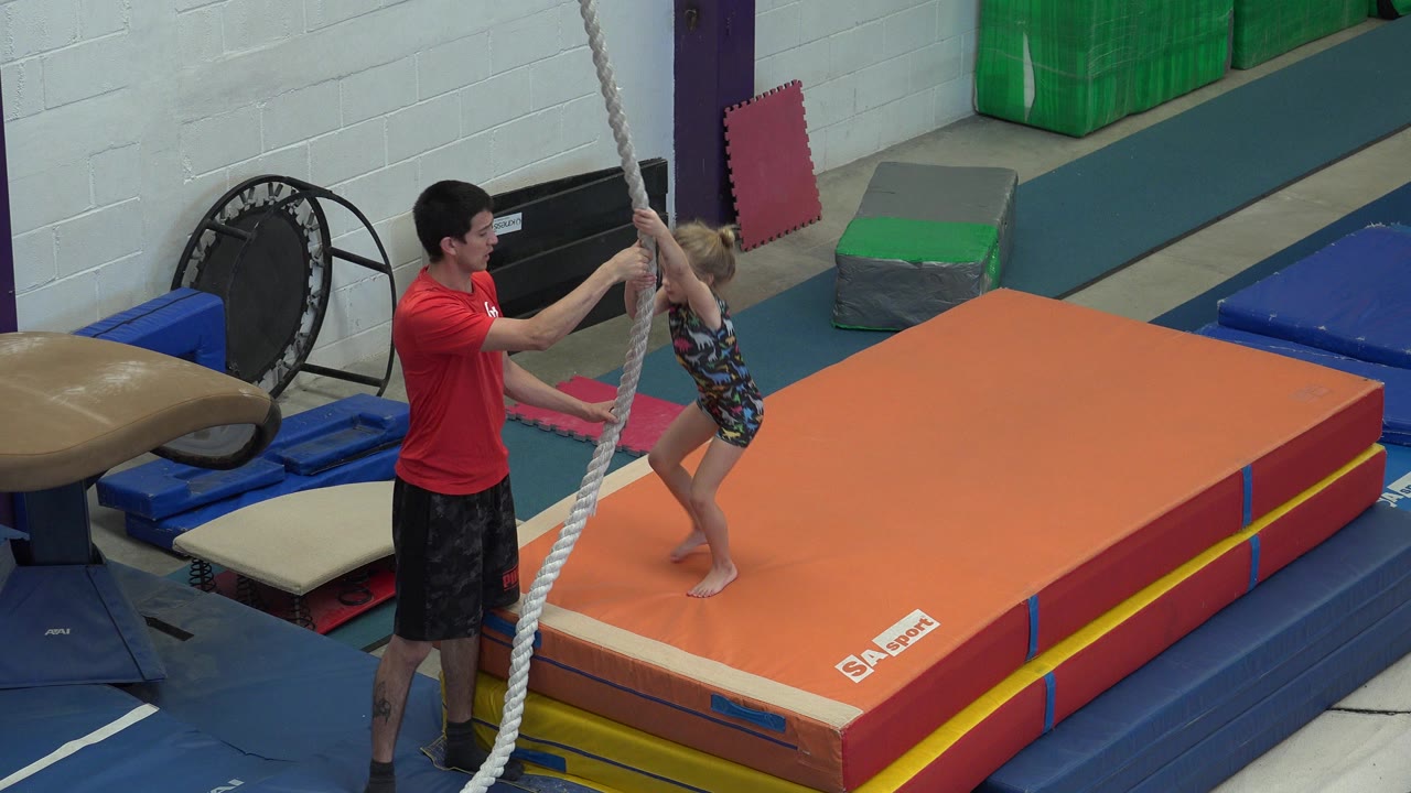Adult man helps a blond girl grip a rope beside an orange gym mat.