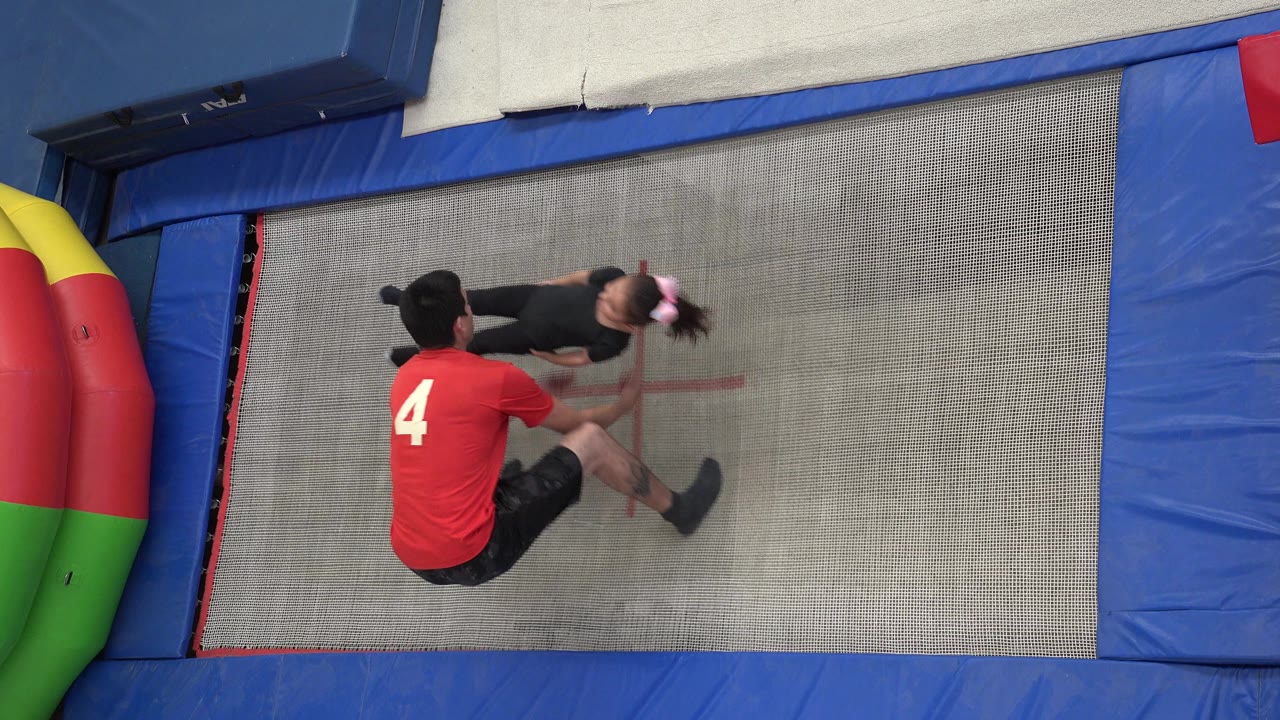 A high overhead view shows the spotter seated or crouched on the trampoline while the girl is held horizontally near his arms.