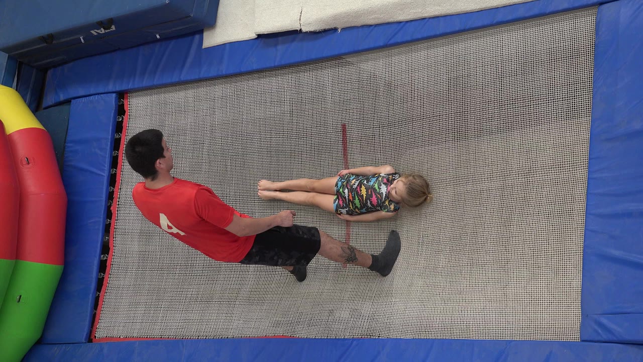 Trainer stands beside a girl lying on a trampoline.