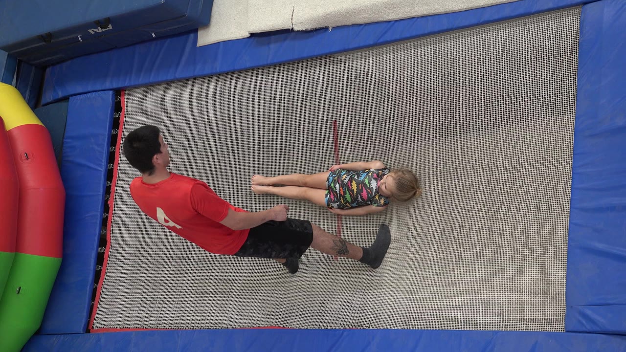 In Cabo San Lucas, Mexico, the trainer stands near the girl's feet while she lies on the trampoline, with the overhead view showing the mat texture and blue padded border.