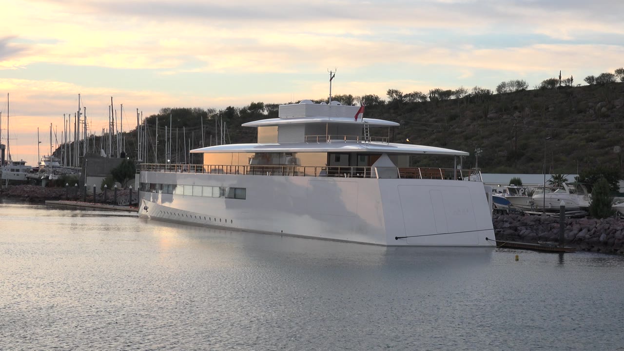 The white tour boat remains docked in Cabo San Lucas, with the bow near the right side of frame, marina masts clustered to the left, muted sunset color in the clouds, and no visible people.