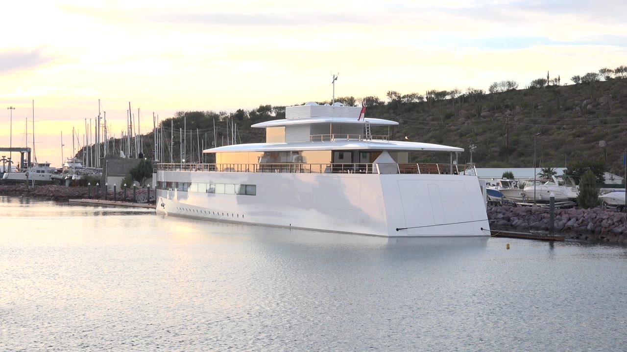 A white three-level mega yacht is moored in a Cabo San Lucas marina with calm water in front, sailboat masts behind, low hillside terrain, and no visible people.