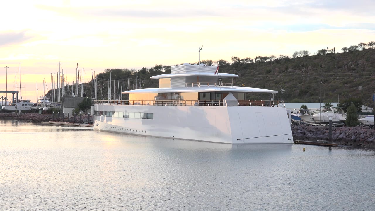 White three-level yacht moored near a rocky marina edge.