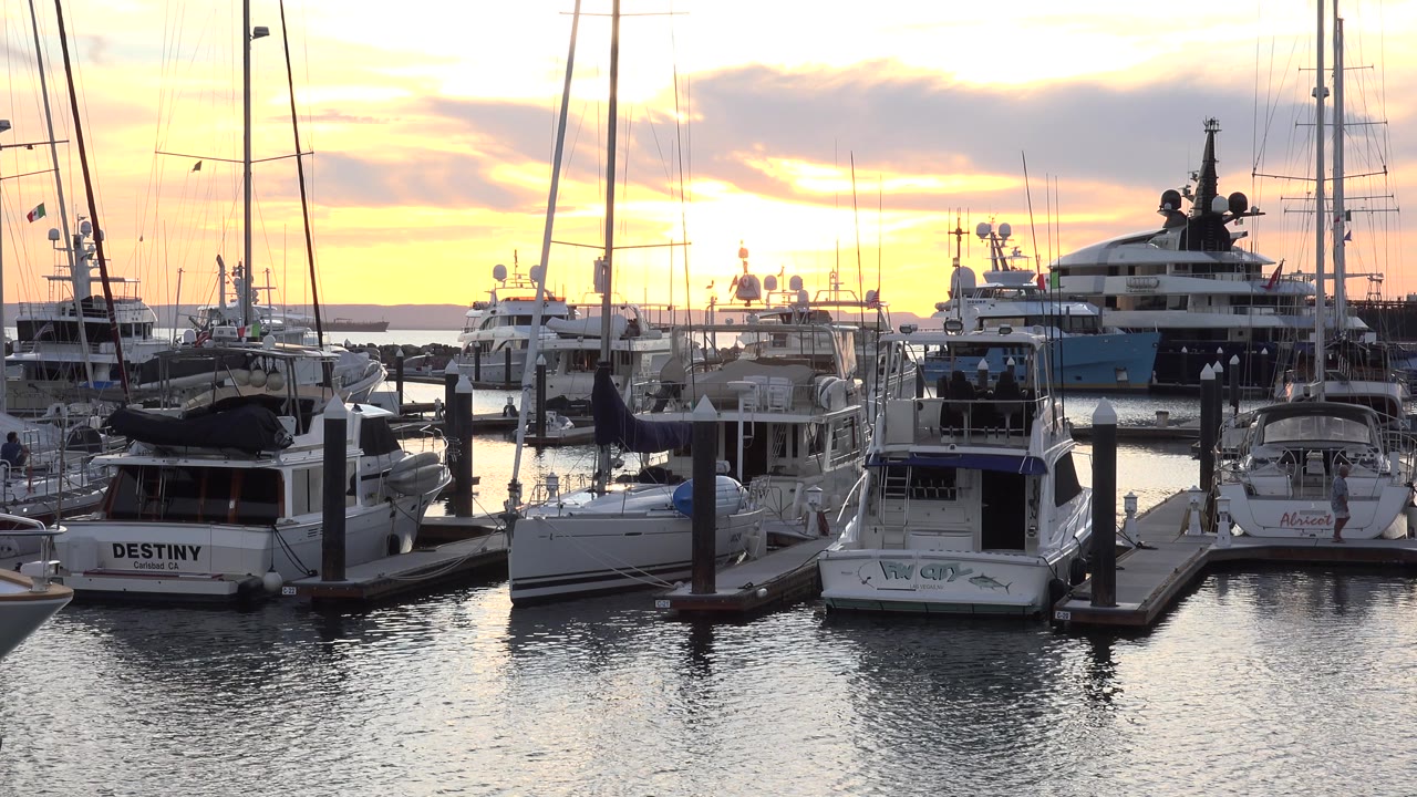 Cabo San Lucas marina frame at 0:01 showing clustered docked boats, upright masts, pier pilings, a large yacht in back, and no visible people.