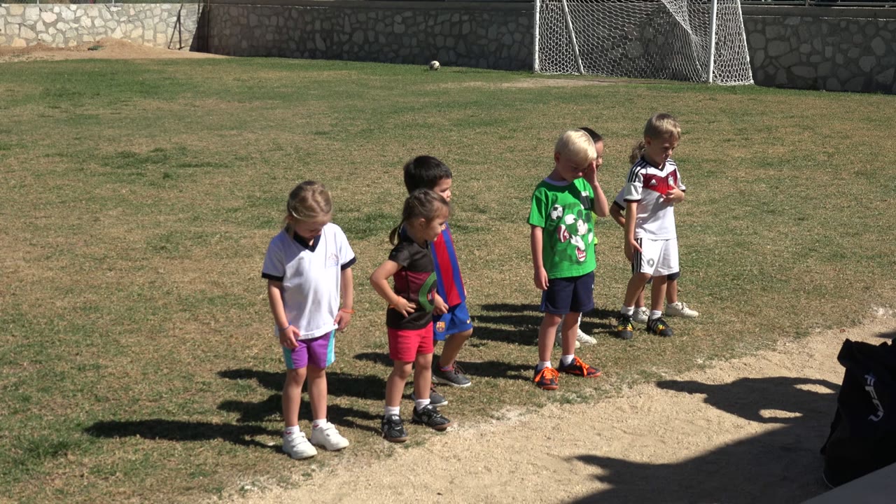A group of children in bright sports shirts stands on a sunlit football field in Los Cabos, Mexico, with goal nets and a stone wall behind them.