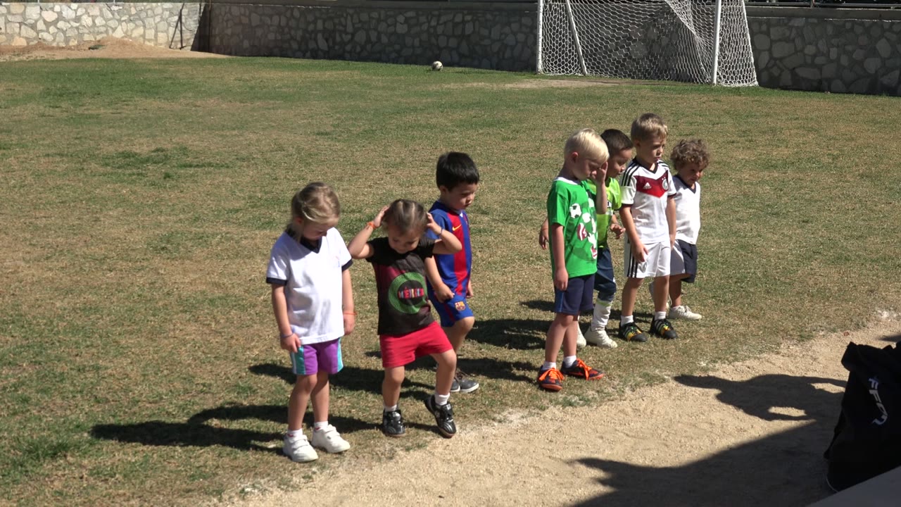 Several children begin to shift their stance on the Los Cabos football field, with one child in red shorts near the front and goal nets in the background.