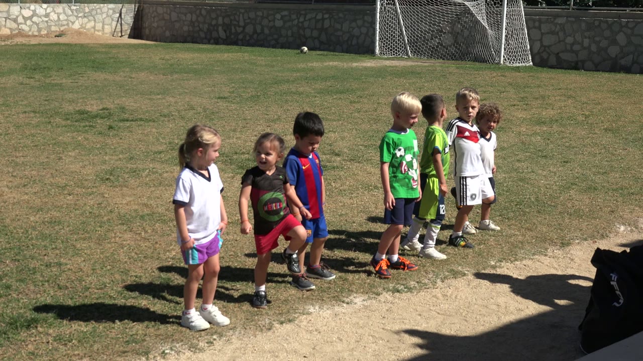 The children remain grouped on the grass in Los Cabos, Mexico, with one child in red shorts stepping forward near the dirt patch.