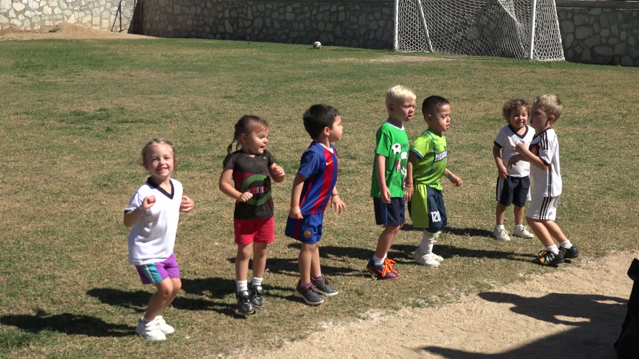 A group of children bounce in place on the Los Cabos grass field, with one child in a white shirt smiling toward the camera.