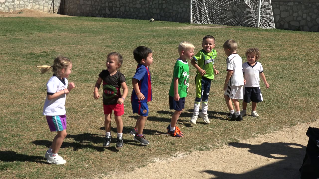 Children move loosely across the grass soccer field in Los Cabos, Mexico, with one girl in a white shirt stepping forward and several boys grouped near the goal area.